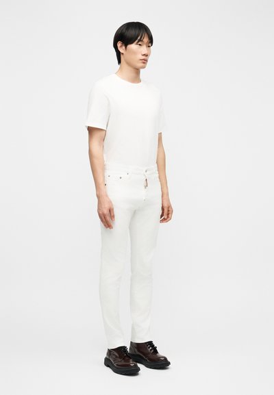 Homme debout de face portant un t-shirt blanc à manches courtes, un pantalon blanc et des chaussures à lacets marron foncé sur un fond blanc.