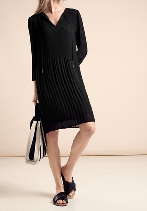 Vrouw in een knielange zwarte geplooide jurk met lange mouwen, draagt een zwart-witte draagtas en zwarte open-toe slippers.