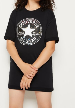 Niño con una camiseta negra extragrande de Converse All Star con logo moteado multicolor, de pie con las manos entrelazadas delante.