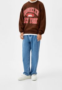Brun sweatshirt med "BROOKLYN NEW YORK" i röd text, kombinerad med ljusblå löst sittande jeans och vita sneakers.