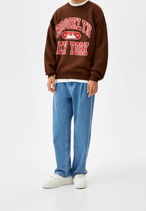 Sudadera marrón con "BROOKLYN NEW YORK" en impresión roja, combinada con vaqueros sueltos de color azul claro y zapatillas blancas.