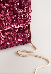 Pochette recouverte de sequins brillants rose foncé avec une bandoulière chaîne en métal doré sur un fond clair uni.