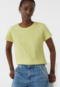 Jasnożółta, teksturowana koszulka z okrągłym dekoltem i krótkimi rękawami, w zestawieniu z niebieskimi dżinsami. Minimalistyczny design, bez widocznych wzorów czy akcentów.