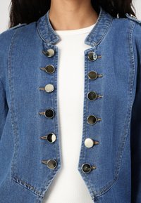 Denim jacket featuring a collared design, button closure with metallic accents, and contrasting fabric underneath. Classic blue color.