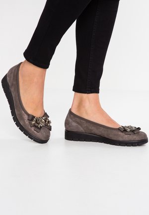 Pieds portant des chaussures compensées basses en daim gris avec des semelles en caoutchouc noir et des nœuds décoratifs ornés de bijoux sur le dessus, associées à un pantalon noir ajusté.