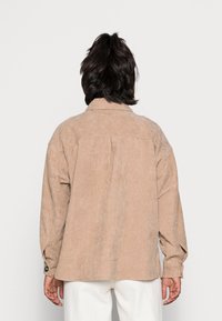 Beiges Cordhemd mit lockerer Passform, langen Ärmeln, spitzem Kragen und Knopfdetails an den Manschetten. Sichtbare gerippte Struktur im Stoff.