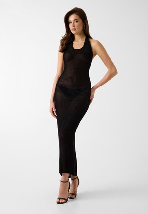 Femme debout dans une robe longue noire sans manches en maille transparente, avec des sous-vêtements noirs et des talons noirs à bride cheville sur un fond uni.