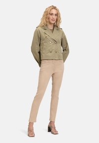 Veste croisée olive avec boutons dorés et deux poches avant, associée à un pantalon slim beige clair et des sandales à talons.