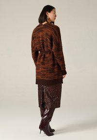 Cardigan a maglia a strisce marroni e arancioni con cintura, indossato sopra una gonna bordeaux con paillettes e stivali lucidi marroni al ginocchio.