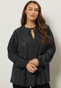 Sort bluse med en nøglehuludskæring med bånd, detaljeret med intrikate spidser og perler, lange ærmer og en afslappet silhuet.