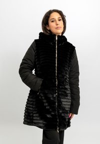 Zwarte textuurjas met een bovenste gedeelte dat op bont lijkt en slanke zwarte mouwen. Voorzien van een ritssluiting aan de voorkant en een capuchon.