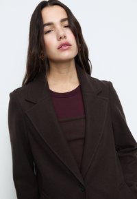 Hnědý vlněný blejzr s dlouhými rukávy, klopami s výstřihem a jedním knoflíkovým zapínáním. Pod ním je vínově červený přiléhavý top.