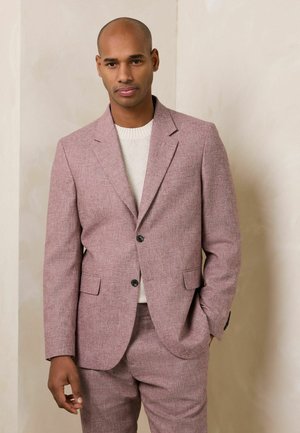 Mann trägt einen hellrosa strukturierten Anzug und einen weißen Strickpullover, steht mit einer Hand in der Tasche vor einem schlichten beigen Hintergrund.