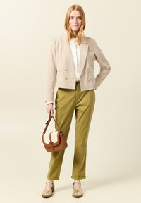 Blazer beige tagliato sopra una camicia bianca, abbinato a pantaloni verde oliva, borsa marrone e scarpe con zeppa beige. Texture lisce e vestibilità sartoriale.