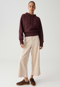 Donna che indossa un hoodie borgogna, pantaloni beige a gamba larga e sneaker marroni, in piedi contro uno sfondo grigio chiaro semplice.