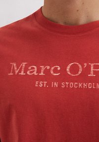 Sarkana kokvilnas T-krekls ar apaļu kakla izgriezumu un nodilušu baltu druku ar tekstu "Marc O'Polo EST. IN STOCKHOLM."