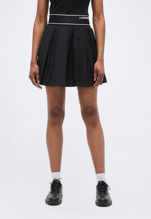 Woman wearing a black pleated skirt with a branded waistband, black lace-up shoes, white socks, and a black sleeveless top.