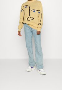 Person som bär ljusblå jeans, vita sneakers med lila detaljer, och en beige tröja med abstrakt ansiktslinjekonst med blå ögon.