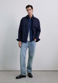 Cămașă cu nasturi de marină, purtată peste o jachetă din denim albastru, asociată cu blugi albaștri deschiși și pantofi negri. Țesătură netedă, croială lejeră și design simplu.