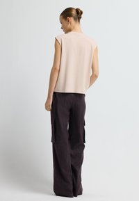 Frau mit einem Dutt, die ein ärmelloses beiges Top und lockere, dunkle Cargo-Hosen trägt, steht mit dem Rücken zu einem schlichten weißen Hintergrund.