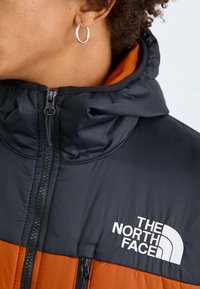 Zblizenie na osobę noszącą czarną i pomarańczową kurtkę North Face z zamkiem błyskawicznym, z widoczną srebrną kolczykiem w kształcie kółka.