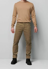 Beżowa koszula z długim rękawem zestawiona z oliwkowozielonymi spodniami i czarnymi sznurowanymi butami. Materiał wydaje się miękki; spodnie mają przednie kieszenie i dopasowany fason.