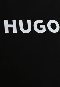 Camiseta negra con un bold texto blanco que dice "HUGO" en una fuente moderna. La tela tiene una textura suave.