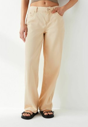 Beige wijde denim met een knoop- en ritsluiting, vijf zakken en zichtbare stikseldetails. Gestyled met zwarte teenslippers.