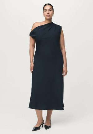 Femme portant une robe midi noire à une épaule et des escarpins noirs à bout pointu, se tenant devant un fond clair.