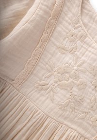 Šviesiai smėlinė medvilninė suknelė su siuvinėtomis gėlėmis. Yra tekstūruotas liemuo, klostuota sijonė ir delikatūs nėriniai aplink iškirpę.