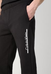 Crne pamučne jogger hlače s bočnim džepovima i bijelim "Calvin Klein" logom na lijevoj nozi. Jednostavan dizajn s udobnim fitom.