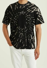 Muž v krátkém rukávovém tričku s černo-bílým tie-dye vzorem a malým bílým textem na hrudi, s bílými kalhotami a jednou rukou v kapse.