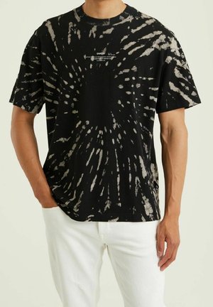 Man wearing black and white tie-dye short sleeve T-shirt with small white text on chest and white pants, one hand in pocket.