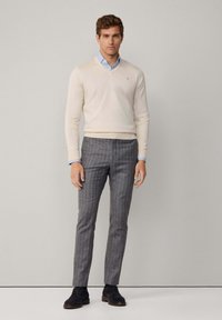 Suéter v-neck beige claro, camisa de cuello azul debajo, combinado con pantalones grises de rayas y zapatos oscuros. Corte sencillo y ajustado.