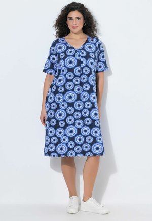 Kvinde iført en knælængde blå kjole med et cirkulært hvidt mønster, korte ærmer og hvide sneakers, står mod en hvid baggrund.