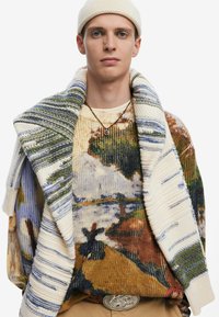 Maglione lavorato a maglia con un motivo paesaggistico testurizzato in tonalità terrose e dettagli crema, sovrapposto a un poncho a righe multicolori.