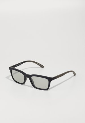 Óculos de sol retangulares com armação preta mate e lentes cinzentas, apresentando realces castanho-esverdeados nas hastes e detalhes em metal.