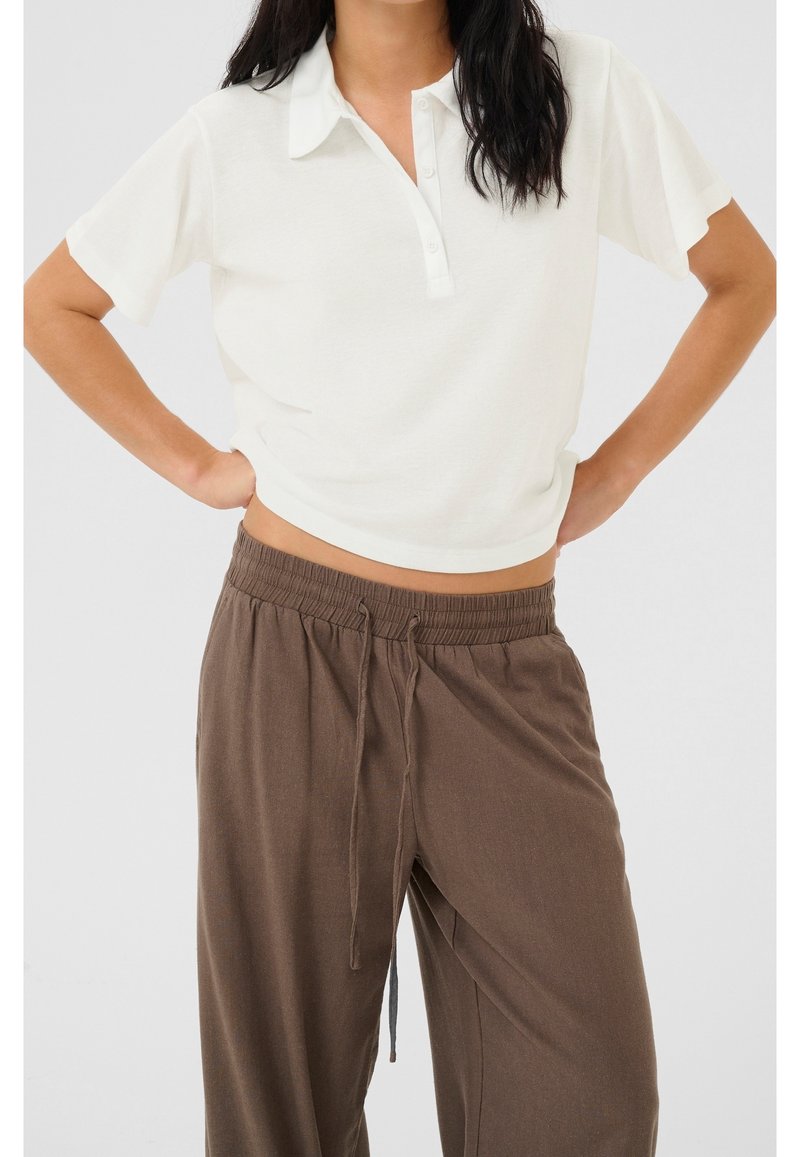 Femme portant une chemise blanche à manches courtes avec col et un pantalon marron ample à cordon, les mains posées sur les hanches.