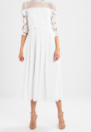 Witte midi-jurk met een kanten lijfje met driekwartmouwen, een vloeiende rok en een lichtgewicht textuur. Gecombineerd met sandalen met bandjes in een nudekleur.