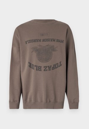 Persoon draagt een oversized bruine sweatshirt met zwarte tekst, zwarte broek, zwarte schoenen, en houdt in één hand een blauwgrijze doek vast.