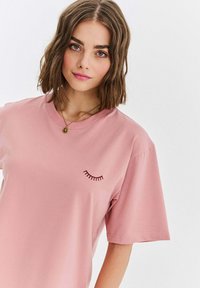 Růžové bavlněné tričko s krátkými rukávy, zdobené vyšívaným motivem řas na levé straně. Hladká textura a volný střih.