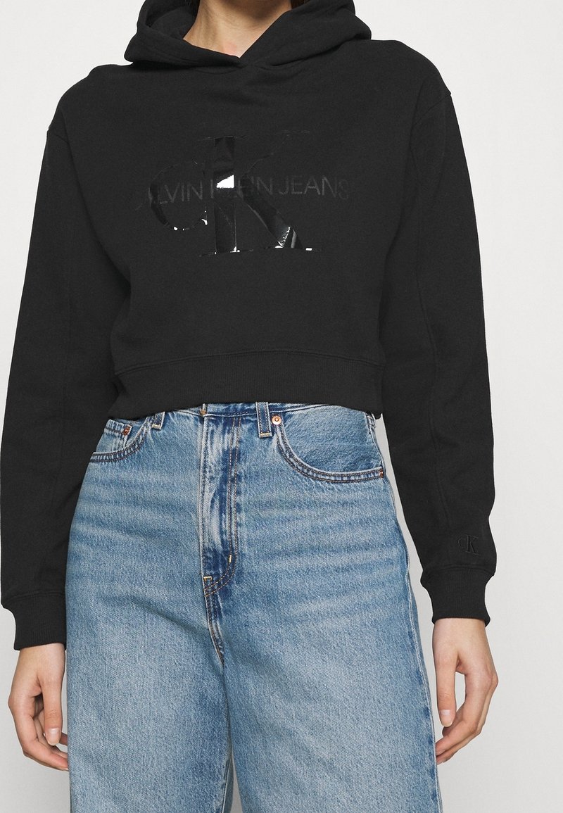 Felpa nera corta con un logo che presenta dettagli riflettenti. Abbinata a jeans chiari a vita alta, per un look casual.