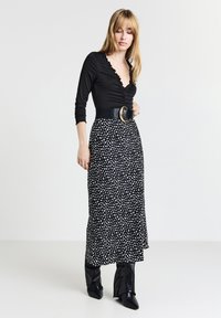 Donna con lunghi capelli biondi che indossa un top nero a scollo a V arricciato, una gonna midi a pois bianchi e neri, una cintura larga e stivali neri con tacco.