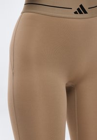 Gros plan sur des leggings taille haute beige avec le logo Adidas noir et une bande noire sur la ceinture, montrant un tissu lisse et des coutures soignées.