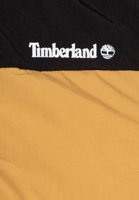 Czarny i musztardowo-żółty materiał tekstylny z białym logo „Timberland” w formie haftu. Charakteryzuje się gładką, trwałą tkaniną z dwukolorowym wzorem.