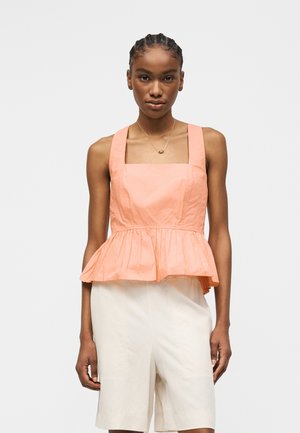 Nuori nainen punotut hiukset, hänellä on päällään hihaton persikanvärinen peplum-toppi ja korkeavyötäröiset valkoiset shortsit yksiväristä taustaa vasten.