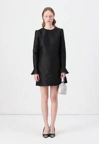 Vestito nero testurizzato con maniche lunghe, polsini svasati e silhouette aderente. La modella tiene in mano una piccola borsa argentata e indossa tacchi neri.