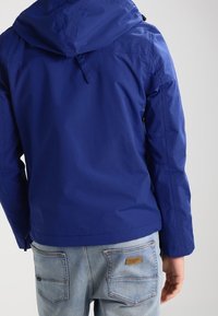 Blå, vandtæt jakke med hætte, der har en glat tekstur og elastiske manchetter. Sammen med lyseblå jeans med baglommer.