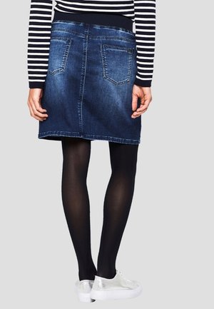 Jupe en denim bleu foncé, avec une taille extensible, des poches arrière standards et une coupe droite. Portée avec des collants noirs et des chaussures argentées.
