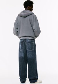 Siva oversized dukserica s kapuljačom, izblijedjeli plavi široki traperice s tamnim pranjem, s velikim stražnjim džepovima i opuštenim krojem. Bijele tenisice.